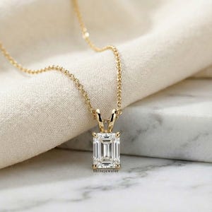 May include: A gold-colored necklace with a rectangular, clear gemstone pendant. The pendant is set in a gold-colored frame and hangs from a delicate chain. The text "ShineJeweler" is visible on the pendant.