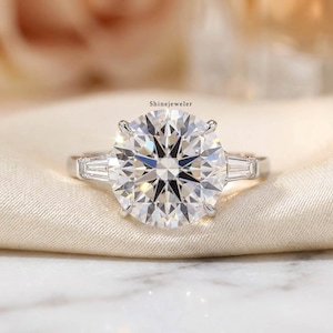 May include: A close-up of a diamond ring with a large, round-cut center stone and two smaller baguette-cut side stones. The ring is set in a silver band and rests on a cream-colored fabric. The brand name "ShineJeweler" is visible.