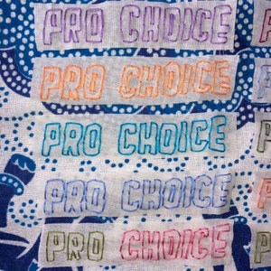 Puede incluir: Cinco parches de tela blanca con las palabras "Pro Choice" bordadas en diferentes colores: morado, naranja, turquesa, azul y rosa.