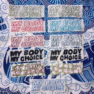 Puede incluir: Primer plano de una tela con varios parches cosidos. Cada parche tiene el texto "MY BODY MY CHOICE" escrito en diferentes colores, incluyendo negro, rojo, verde y azul.