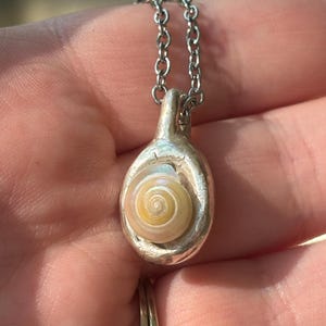 May include: A silver pendant necklace featuring a small, circular, cream and tan spiral seashell set into a textured, organic-shaped silver frame. The pendant hangs from a simple silver chain link, showcasing a natural, iridescent shell center.