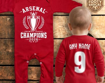 Puede incluir: Body rojo para bebé con texto y gráficos blancos. La parte delantera presenta "ARSENAL" sobre un gráfico de trofeo y "PREMIER LEAGUE CHAMPIONS 2026". La parte trasera tiene "ANY NAME" y el número "9".
