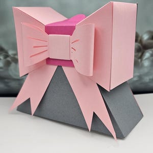 Może przedstawiać: Geometryczne papierowe pudełko na prezent z różową kokardą i szarą podstawą. Kokarda ma wycięte detale i jest ułożona na ciemniejszym różowym środku. Pudełko ma unikalny kształt, idealne na małe prezenty lub do celów dekoracyjnych.