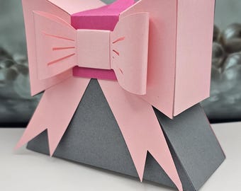 May include: A geometric paper gift box with a pink bow and gray base. The bow has cut-out details and is layered over a darker pink center. The box is a unique shape, perfect for small gifts or decorative purposes.