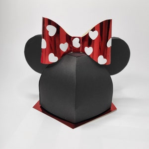 Op de afbeelding: Een zwarte en rode Minnie Mouse-thema cadeaudoos met een rode strik en witte stippen. De doos is gevormd als Minnie Mouse's hoofd met grote oren.