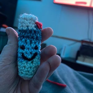 May include: A small crocheted blue and white pill bottle with a red cap and black eyes. The bottle has a smiling face.