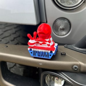 May include: A red, white, and blue 3D printed Jeep Wrangler toy with the words "Jeep Duck" printed on the front. The toy is sitting on the front bumper of a silver Jeep Wrangler.