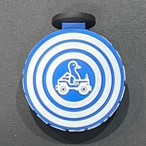 May include: A blue and white round bottle stopper with a black handle. The stopper features a duck driving a jeep in a blue circle, surrounded by concentric white and blue rings. The product is likely for a bottle.