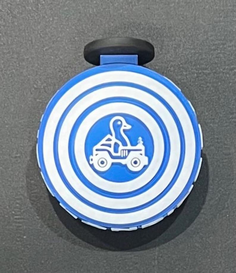 May include: A blue and white round bottle stopper with a black handle. The stopper features a duck driving a jeep in a blue circle, surrounded by concentric white and blue rings. The product is likely for a bottle.