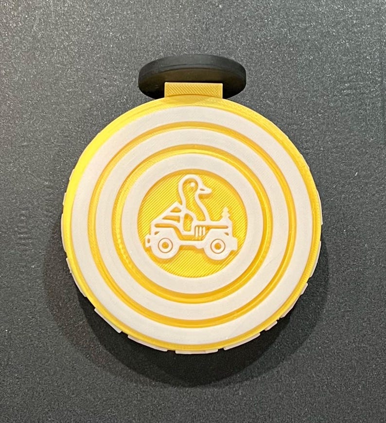 May include: A yellow and white circular pendant with a black top. The pendant features a duck driving a jeep in the centre, surrounded by concentric white circles. The background is a textured yellow.