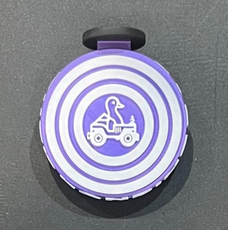 May include: A purple and white bottle stopper featuring a duck driving a jeep. The stopper has a black handle and a circular design with concentric white rings. The duck and jeep are in the centre of the stopper.