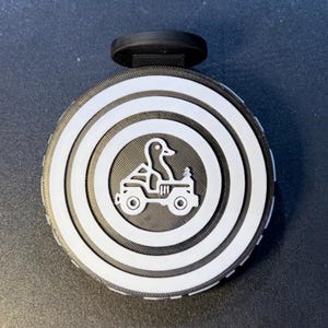 May include: A round bottle stopper featuring a duck driving a jeep, with a black handle. The design has concentric white rings on a black background. The duck graphic is in the centre of the stopper.