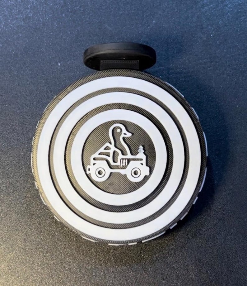 May include: A round bottle stopper featuring a duck driving a jeep, with a black handle. The design has concentric white rings on a black background. The duck graphic is in the centre of the stopper.