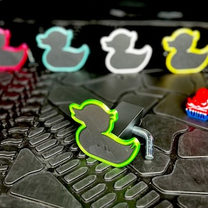 Pu&ograve; includere: Una collezione di ciondoli a forma di anatra colorati. I ciondoli hanno un interno nero e sono bordati in vari colori, tra cui rosa, verde, blu e giallo. Un fermaglio in metallo nero &egrave; attaccato a uno dei ciondoli. Su una superficie nera.