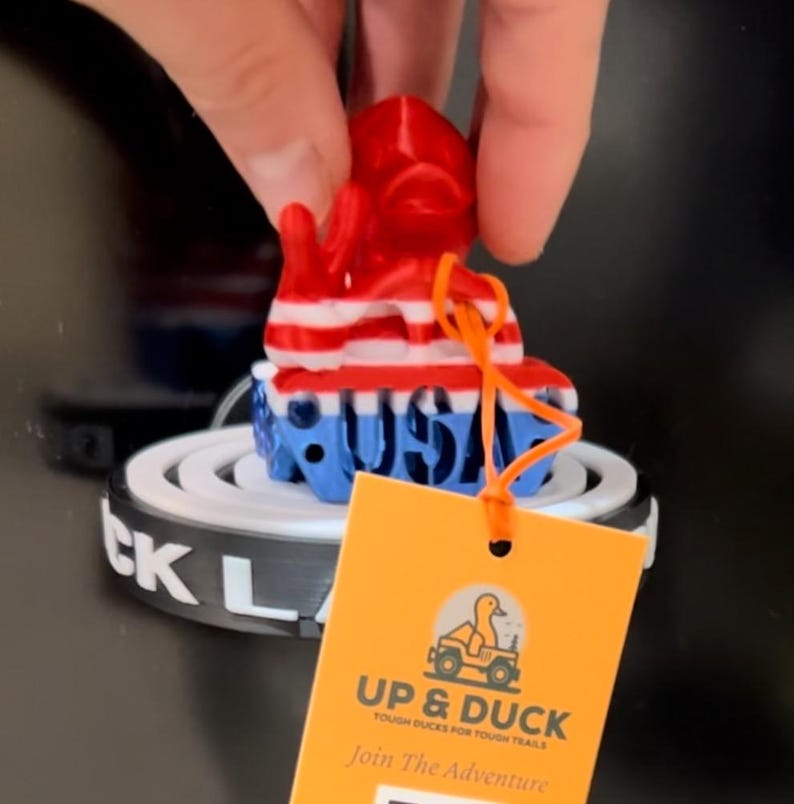 May include: A novelty duck-shaped keyring in red, white, and blue, with "USA" on the base. The keyring is held by a hand and has an orange tag with the text "UP & DUCK" and "Join The Adventure."