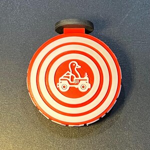 May include: A red and white circular bottle stopper with a duck driving a vehicle design. The design includes concentric circles and a black top. The stopper is likely made of plastic or a similar material.