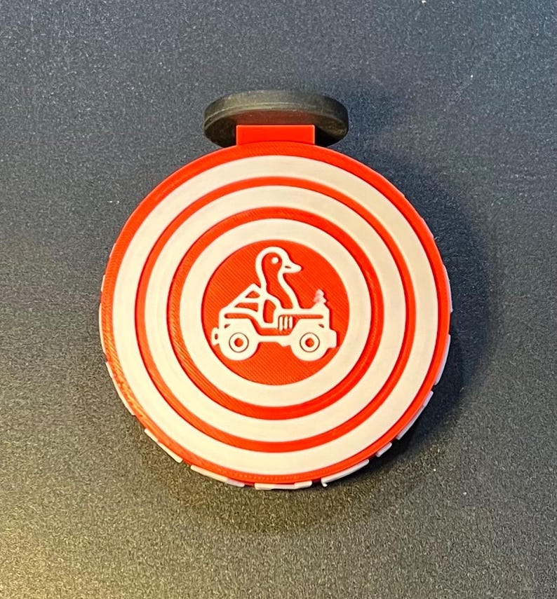 May include: A red and white circular bottle stopper with a duck driving a vehicle design. The design includes concentric circles and a black top. The stopper is likely made of plastic or a similar material.