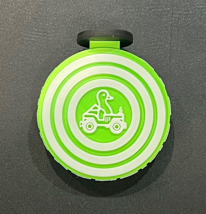 May include: A circular, green and white 3D-printed item with a black clip. It has a concentric circle pattern and a central graphic of a duck driving a jeep. The duck and jeep are green.