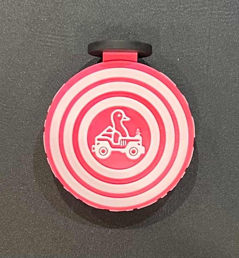 May include: A circular badge with a pink and white target design, featuring a duck driving a jeep. The badge has a black clip at the top. The design is playful and whimsical.