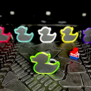 Pu&ograve; includere: Una collezione di ciondoli a forma di anatra in vari colori. I ciondoli sono neri con contorni colorati al neon, tra cui verde, giallo e rosa. &Egrave; presente anche un piccolo ciondolo rosso e blu. I ciondoli sono disposti su una superficie nera strutturata.