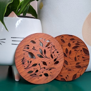 May include: Two round wooden coasters featuring a cut-out floral design. The coasters are brown and have a natural wood finish.