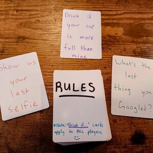 May include: A set of white cards with handwritten rules for a drinking game. The cards include prompts like "Drink if your cup is more full than mine" and "Show us your last selfie". The rules state that "Drink if..." cards apply to all players.