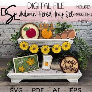 以下が含まれることがあります： 秋をテーマにしたアイテムで飾られた白い段々になったトレイ。赤いリンゴ、カボチャ、ひまわりのガーランド、