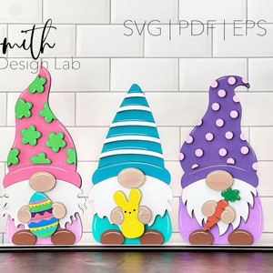 May include: Three wooden gnomes with Easter-themed hats and accessories. The gnome on the left has a pink hat with green shamrocks, a white beard, and is holding an Easter egg. The middle gnome has a blue and white striped hat, a white beard, and is holding a yellow bunny. The gnome on the right has a purple hat with pink polka dots, a white beard, and is holding a carrot.