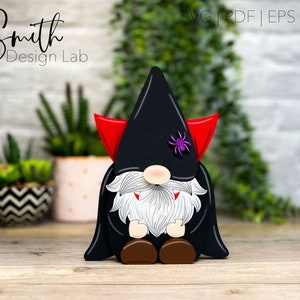 May include: A black and red vampire gnome with a white beard and fangs. The gnome is wearing a black hat with a purple spider on it. The gnome is sitting on a wooden surface.