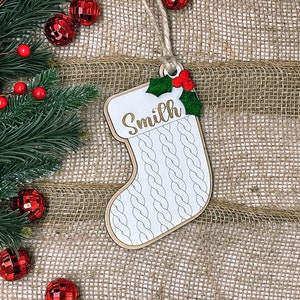 May include: A white wooden Christmas stocking ornament with a knitted pattern and the name "Smith" written in gold lettering. The ornament has a green holly sprig with red berries.