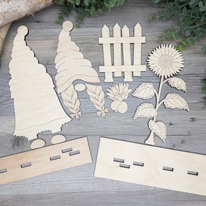 May include: Wooden craft kit featuring two gnomes, a picket fence, a sunflower, and two wooden bases. The kit is designed for painting and decorating.
