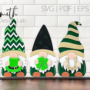 May include: Three gnomes with green hats and white beards. The gnome on the left has a green chevron hat and is holding a green leprechaun hat. The gnome in the middle has a black hat with a shamrock pattern and is holding a green four-leaf clover. The gnome on the right has a green and gold striped hat and is holding a gold horseshoe.
