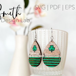 May include: A pair of wooden earrings with a green shamrock design and brown and green striped accents. The earrings are hanging from silver hooks.