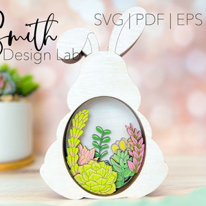 May include: White wooden bunny rabbit cutout with a circular opening in the center. The opening is filled with green and pink succulents. The bunny is standing on a wooden surface.