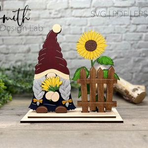 May include: A wooden gnome figurine holding sunflowers, standing next to a wooden fence and sunflower. The gnome is wearing a burgundy hat and a blue outfit. The text "Smith Design Lab" is visible in the top left corner of the image.