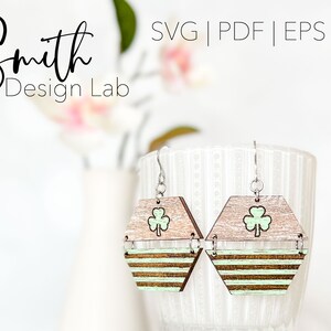 May include: A pair of wooden earrings with a green shamrock design. The earrings are hexagonal in shape and have a striped pattern. The earrings are hanging from silver hooks.