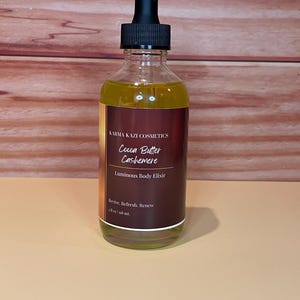 May include: A clear glass bottle of Karma Kazi Cosmetics Cocoa Butter Cashemere Luminous Body Elixir. The bottle has a black dropper top and a brown label with white text. The bottle contains a yellow liquid and is 4 fl oz / 118 ml.