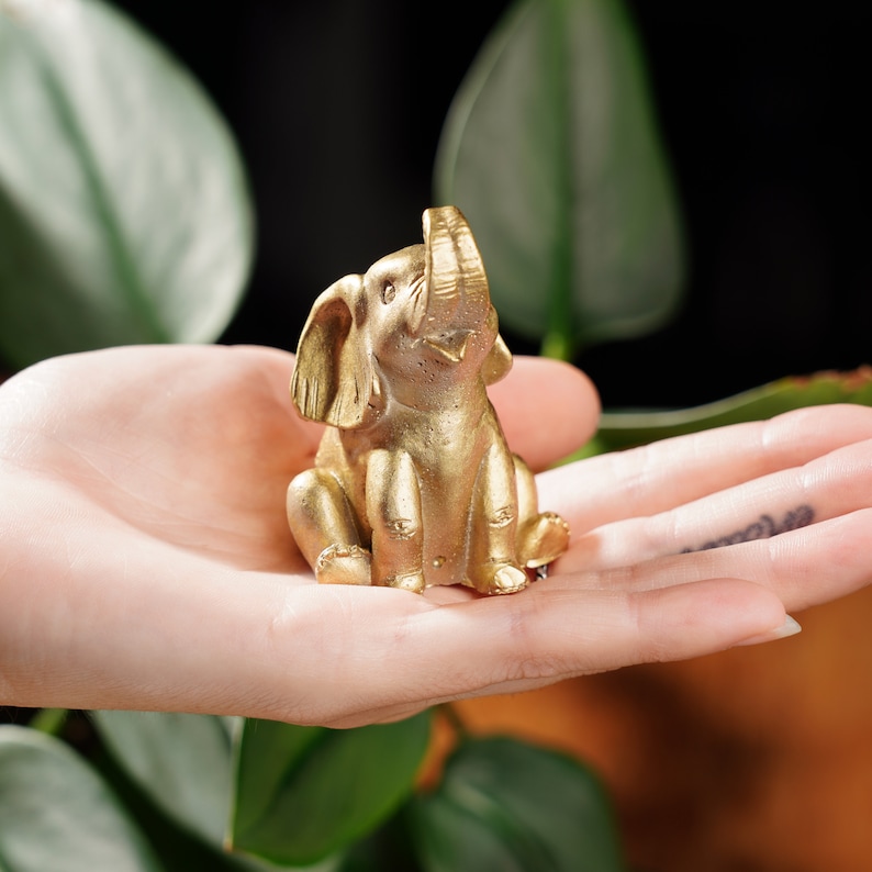 May include: A small, gold-coloured elephant figurine with its trunk raised in the air. The figurine is sitting in the palm of a person's hand.