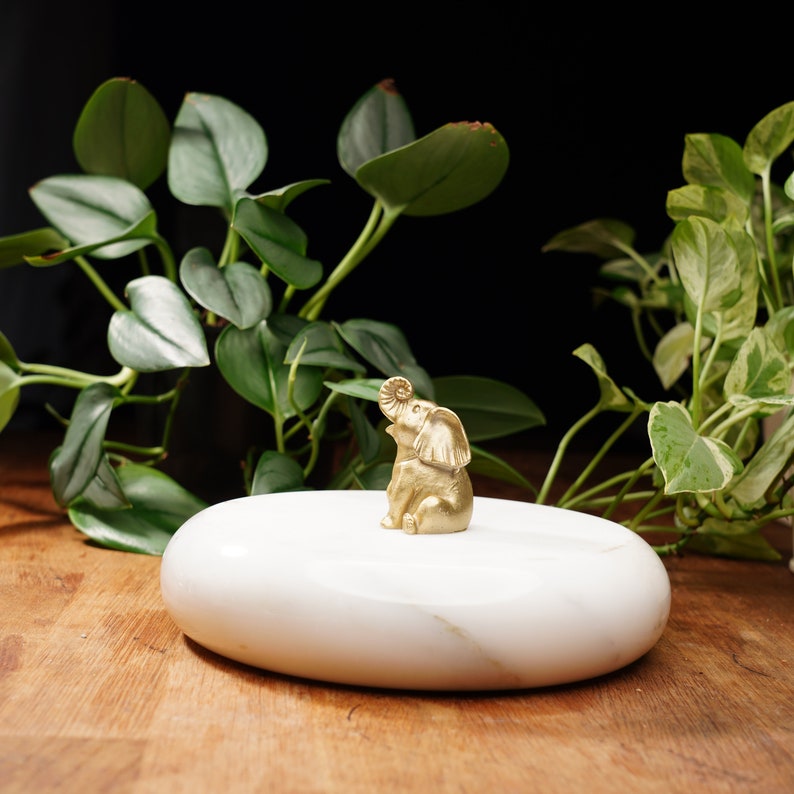 May include: A small gold elephant figurine sits on a white marble oval-shaped dish. The dish is on a wooden surface with green plants in the background.