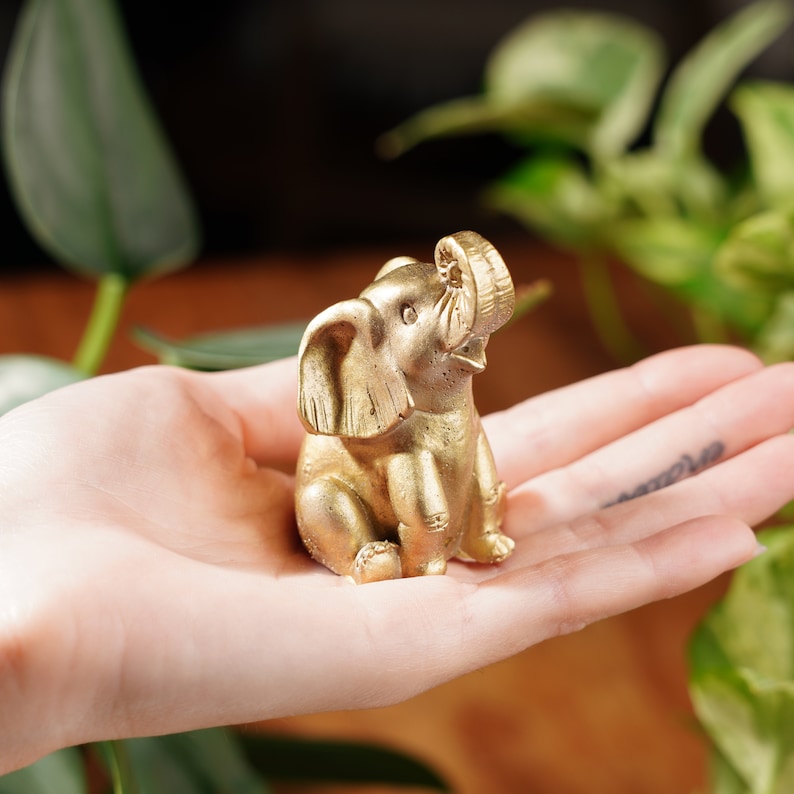 May include: A small, gold-coloured elephant figurine with its trunk raised in the air. The figurine is sitting in the palm of a person's hand.