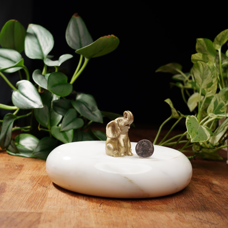 May include: A small gold elephant figurine sitting on a white marble oval-shaped dish. A silver coin is next to the elephant.