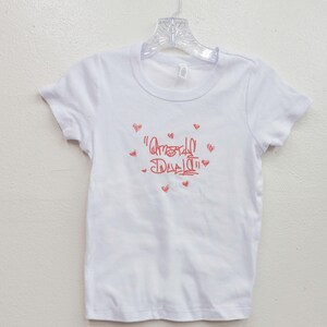 May include: White t-shirt with a crew neck and short sleeves. The shirt features red text and small heart graphics. The text reads "Amor de Dad" in a handwritten style. The shirt is hanging on a clear hanger.