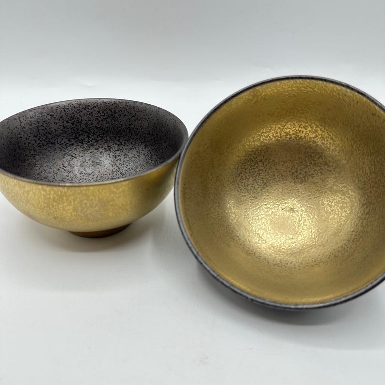 May include: Two decorative bowls with a metallic gold exterior and a speckled black interior. The bowls have a rounded shape and sit on a small base. The interior of one bowl is a solid gold color.