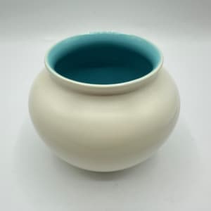 May include: A cream-colored ceramic vase with a rounded body and a wide opening. The interior of the vase is a vibrant teal color, creating a striking contrast with the exterior. The vase has a smooth, glossy finish.