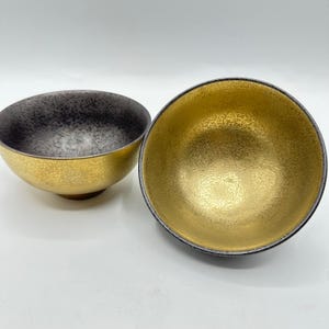 May include: Two decorative bowls with a gold exterior and a contrasting interior. One bowl has a black and gray speckled interior, while the other has a gold interior. The bowls have a metallic sheen and a simple, elegant design.
