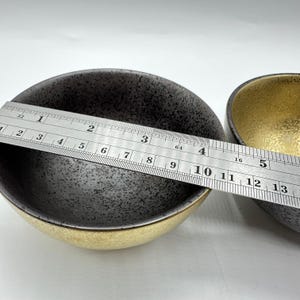 May include: Two small, round ceramic bowls with a speckled black interior and a gold rim. A metal ruler is placed across the bowls, showing measurements. The bowls are likely for serving or decorative purposes.