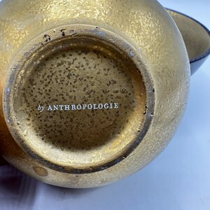 May include: Close-up of a gold-colored decorative bowl with a textured surface. The bottom of the bowl has the text "by ANTHROPOLOGIE" in white. A second bowl is partially visible in the background.