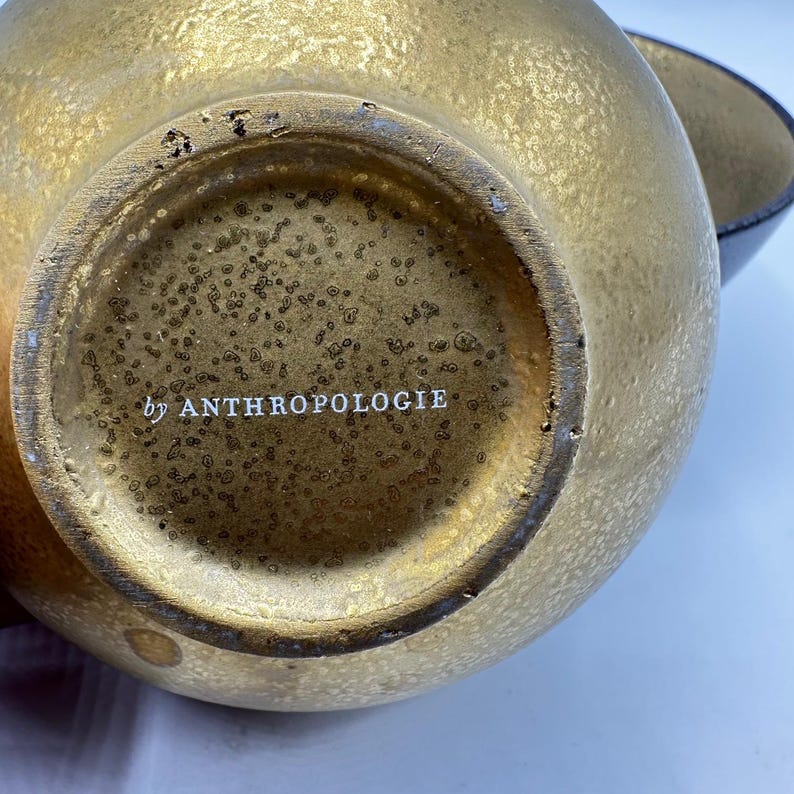May include: Close-up of a gold-colored decorative bowl with a textured surface. The bottom of the bowl has the text "by ANTHROPOLOGIE" in white. A second bowl is partially visible in the background.