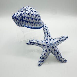 May include: A ceramic seashell and starfish set. The seashell is white with a blue fishnet pattern and a blue rim. The starfish is white with a blue fishnet pattern. The set is displayed on a clear stand.