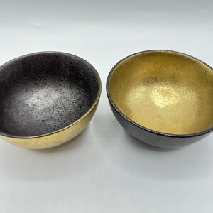 May include: Two decorative bowls with a unique design. One bowl has a black exterior and a gold interior. The other bowl has a gold interior and a black exterior. The bowls have a speckled texture.